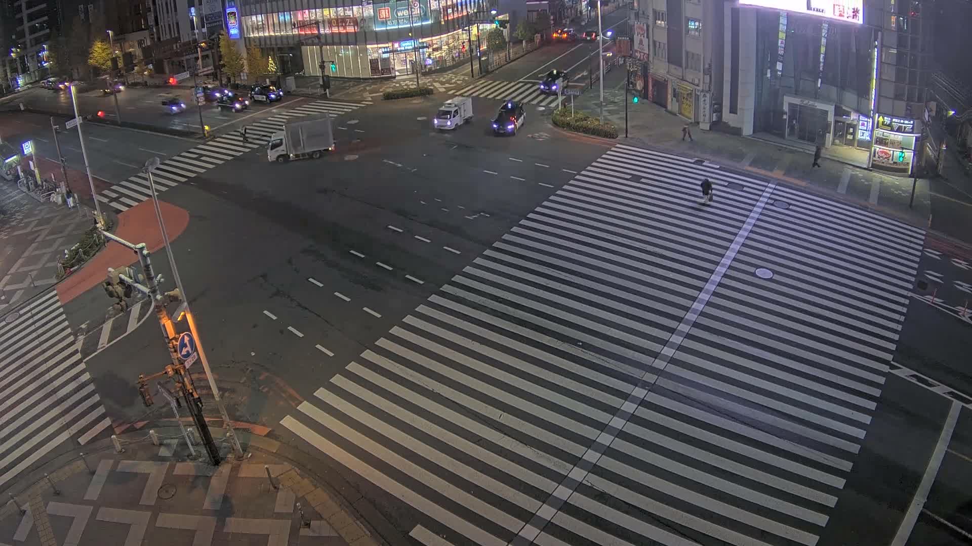 An elevated view shows a bustling city intersection at night, with a dense flow of vehicles including many taxis, and numerous pedestrians crossing well-marked crosswalks between brightly lit buildings under clear, dry weather conditions.