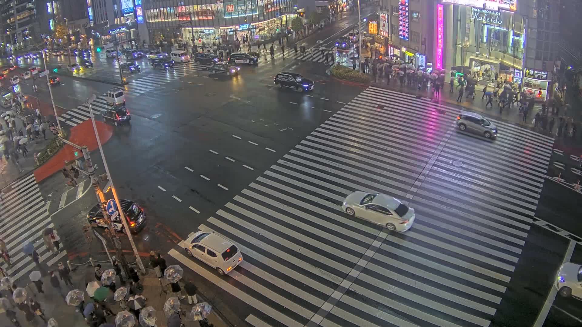 An elevated view shows a bustling city intersection at night, with a dense flow of vehicles including many taxis, and numerous pedestrians crossing well-marked crosswalks between brightly lit buildings under clear, dry weather conditions.