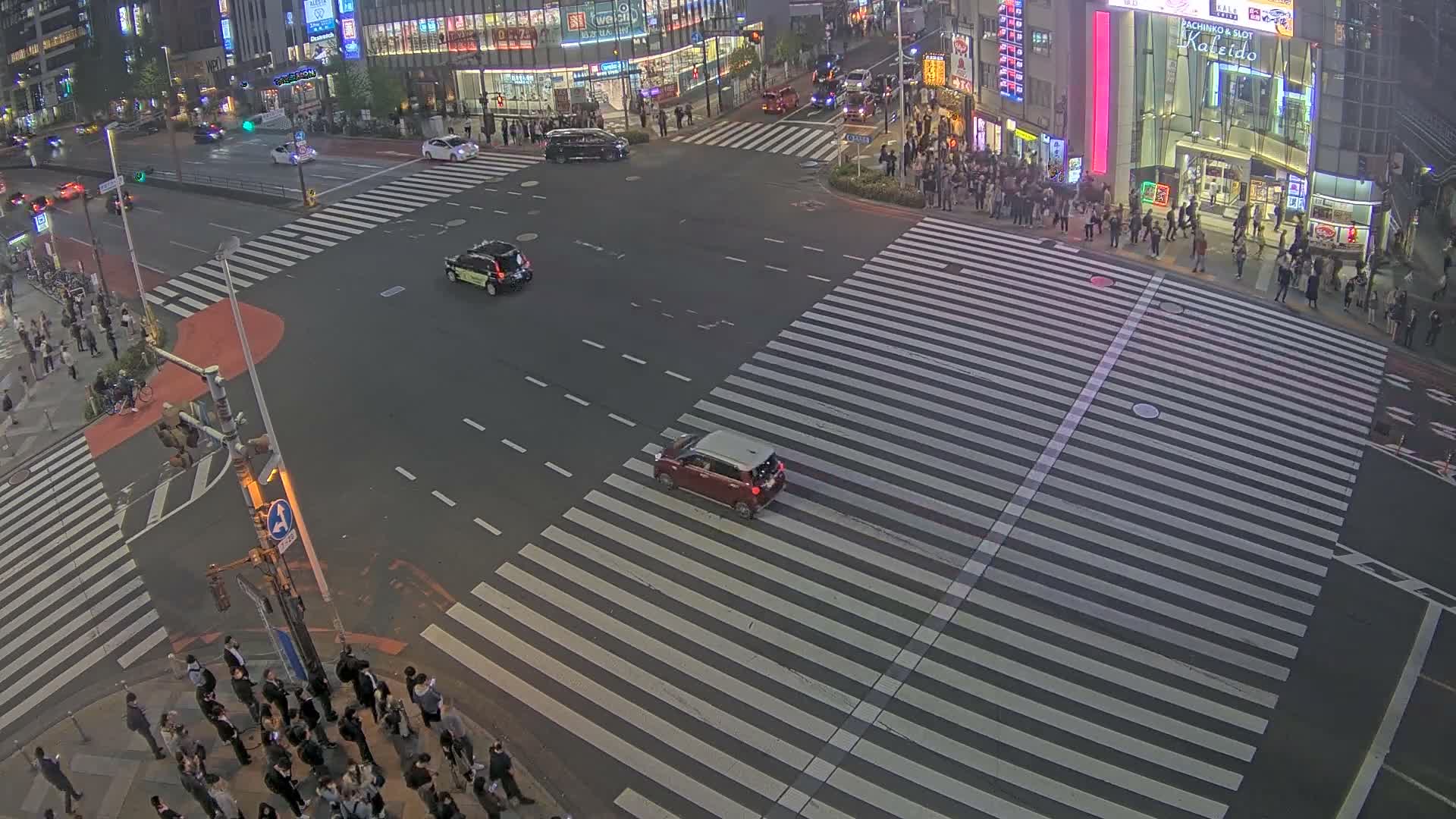 An elevated view shows a bustling city intersection at night, with a dense flow of vehicles including many taxis, and numerous pedestrians crossing well-marked crosswalks between brightly lit buildings under clear, dry weather conditions.
