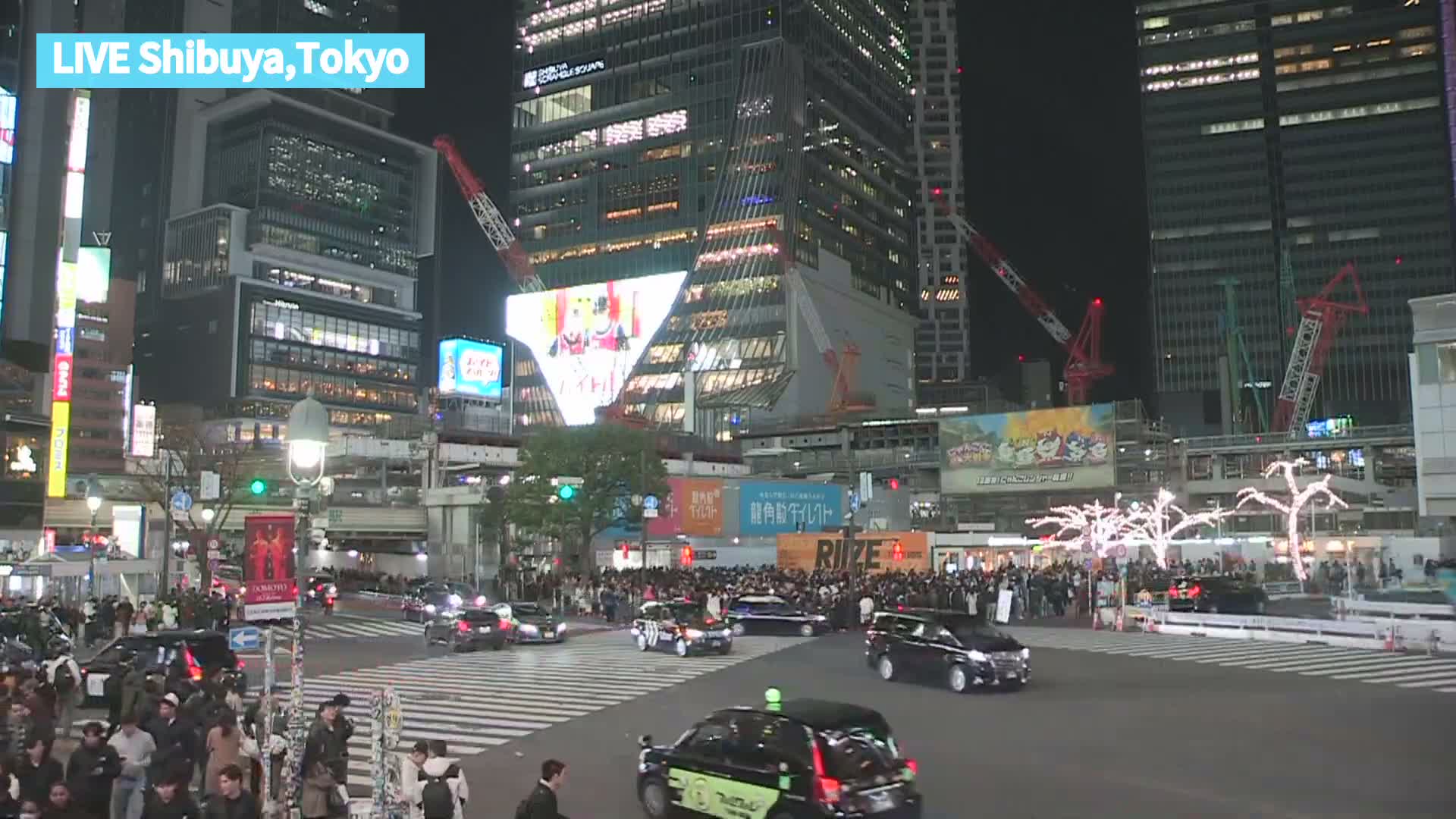 A vibrant nighttime cityscape features numerous pedestrians and cars on a busy street, surrounded by brightly lit skyscrapers and large digital displays, under a clear dark sky.