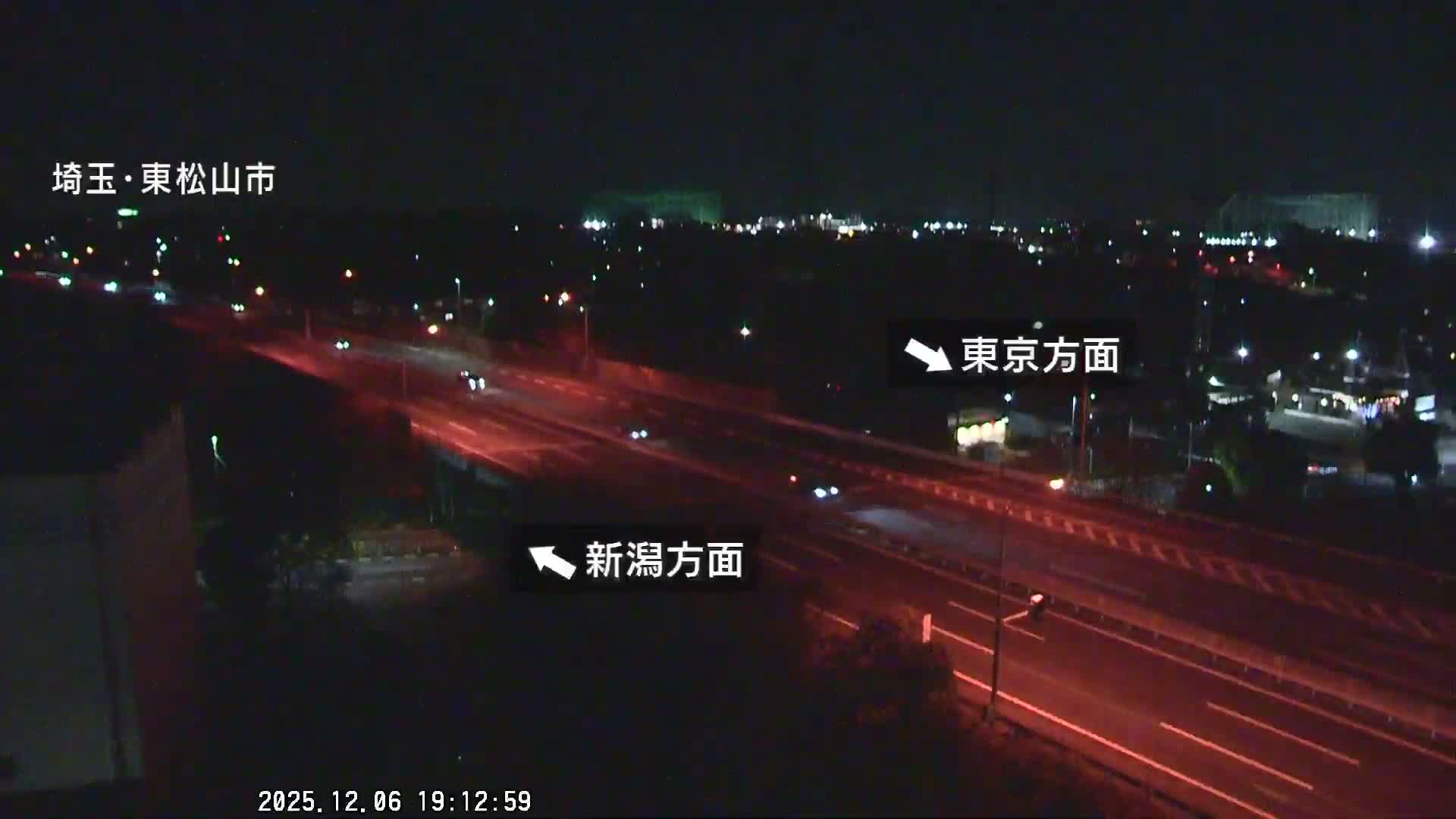 A nighttime view overlooks a multi-lane highway with several cars, their lights visible as streaks and dots, set against a backdrop of distant city lights under a clear sky.