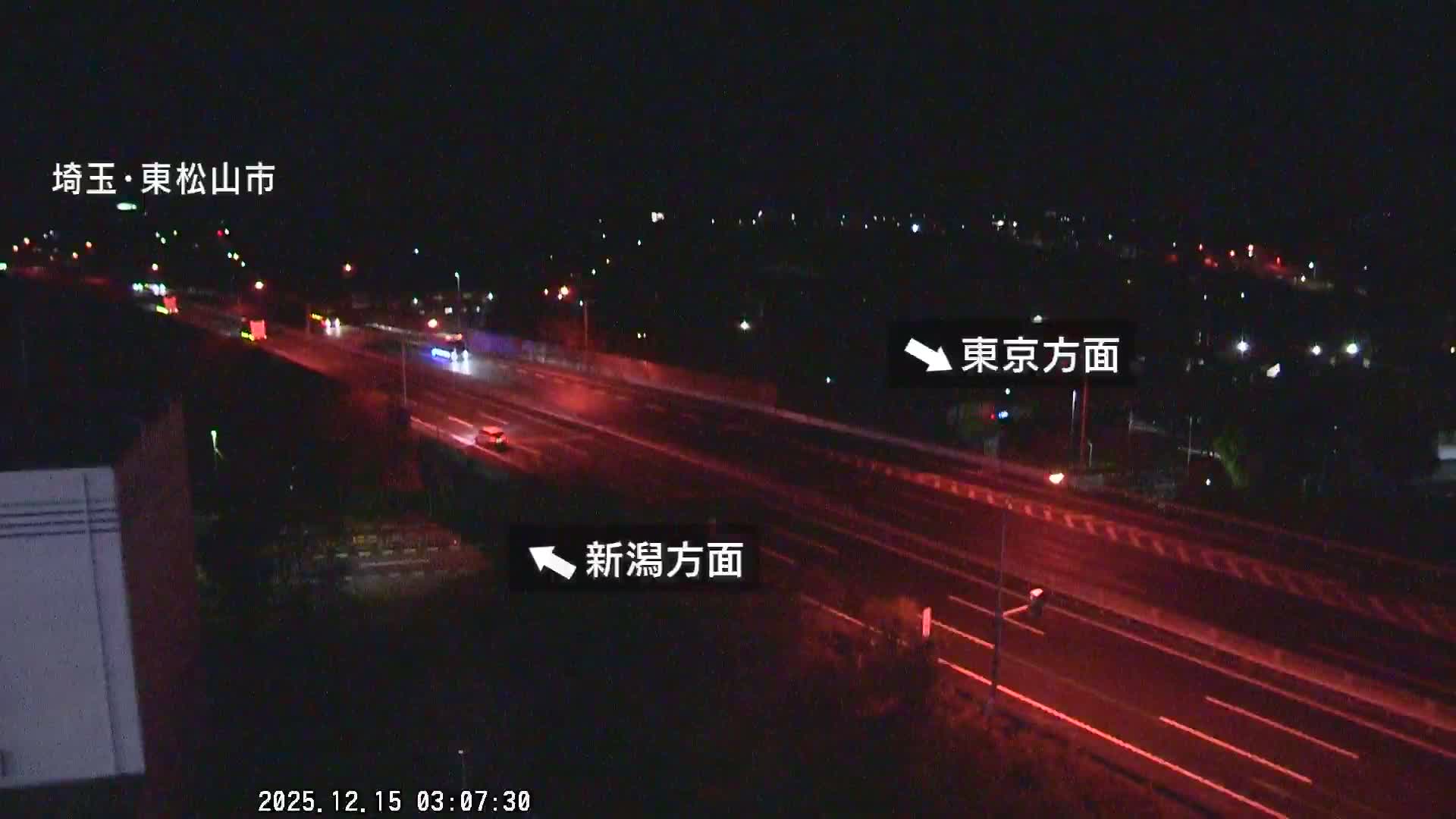 A nighttime view overlooks a multi-lane highway with several cars, their lights visible as streaks and dots, set against a backdrop of distant city lights under a clear sky.