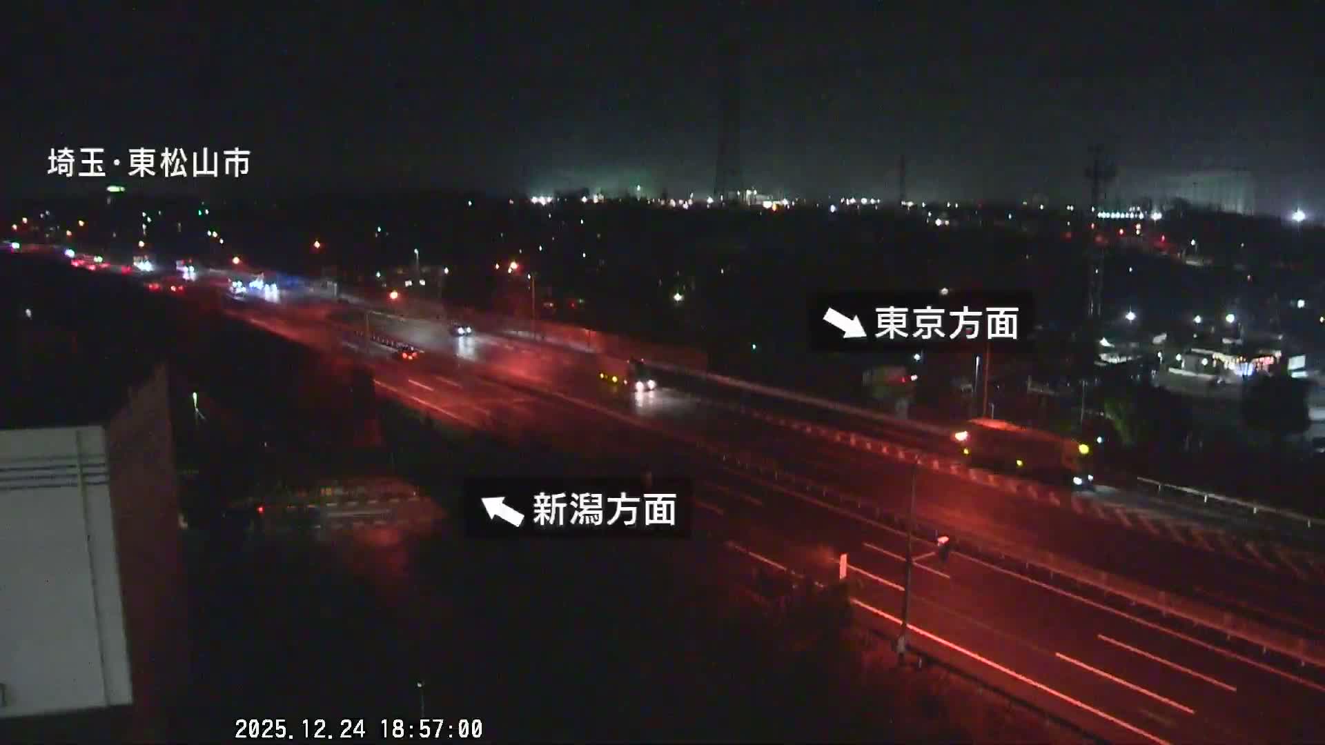 A nighttime view overlooks a multi-lane highway with several cars, their lights visible as streaks and dots, set against a backdrop of distant city lights under a clear sky.