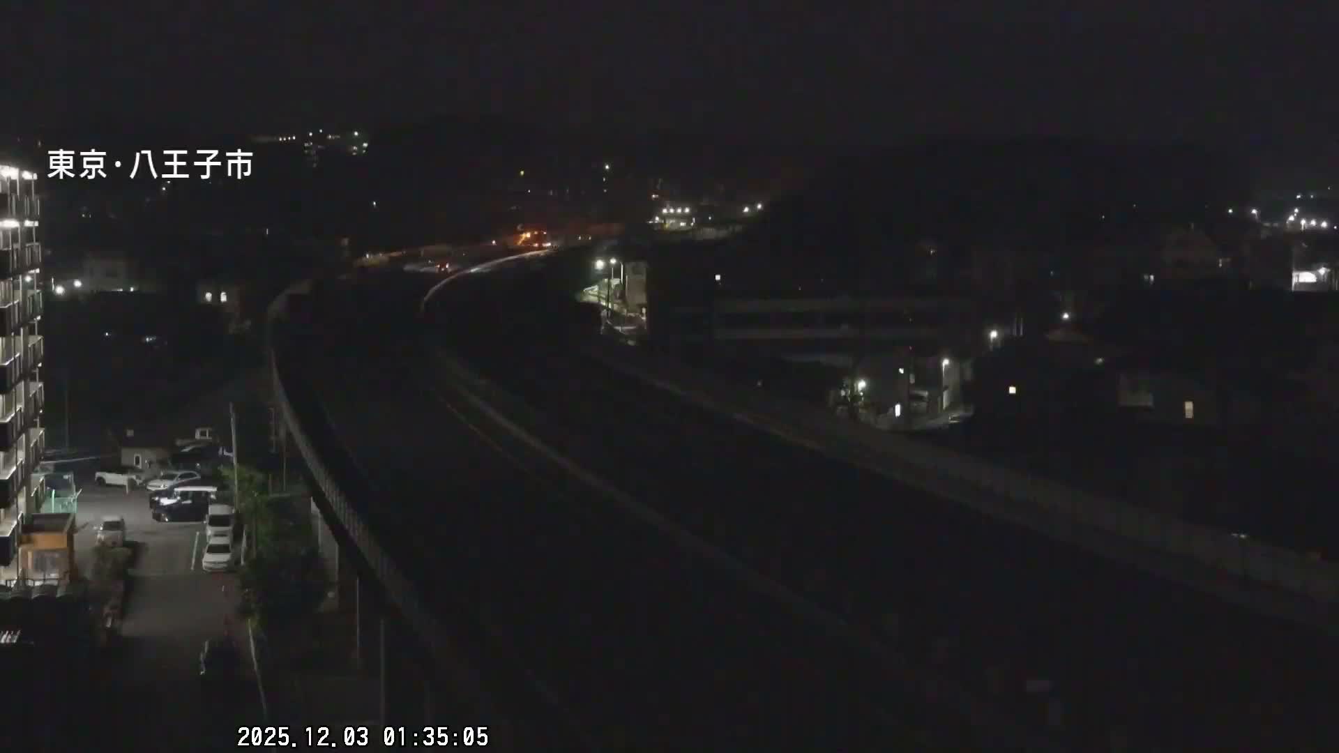 On a clear night, an elevated highway curves over railway tracks, flanked by an apartment building and parked cars to the left, with scattered city lights and dark hills visible in the background.