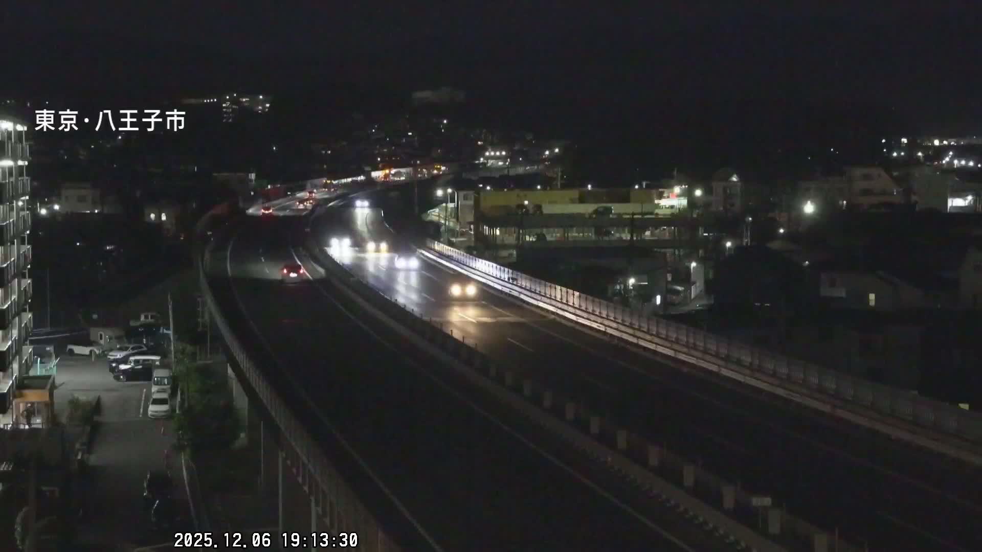 The image displays an elevated highway at night with visible vehicle headlights and taillights, surrounded by illuminated buildings and city lights under clear weather conditions.