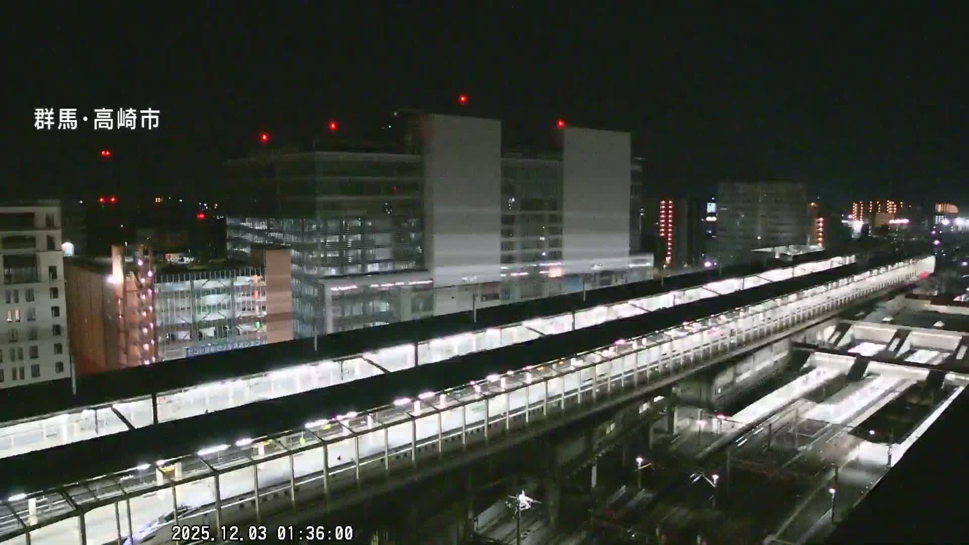 At night, a brightly lit train station with multiple platforms and tracks dominates the foreground, backed by numerous illuminated city buildings under a clear sky.