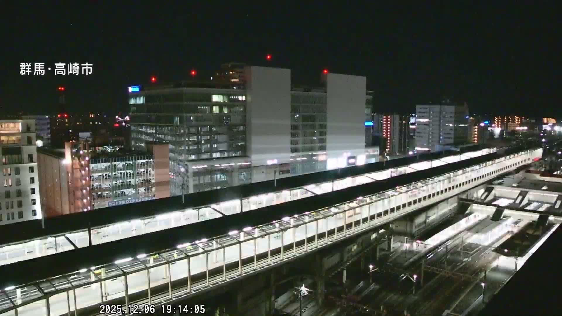 A nighttime aerial view reveals a sprawling city train station with elevated platforms and numerous tracks, flanked by illuminated buildings, all under a clear sky.