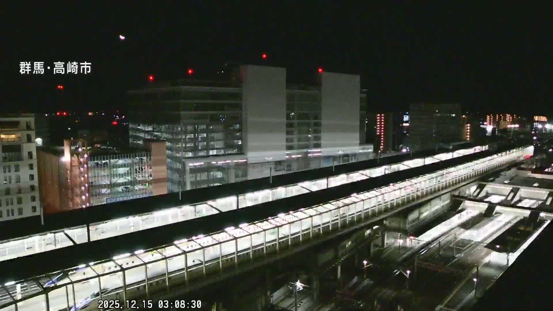 A nighttime aerial view reveals a sprawling city train station with elevated platforms and numerous tracks, flanked by illuminated buildings, all under a clear sky.