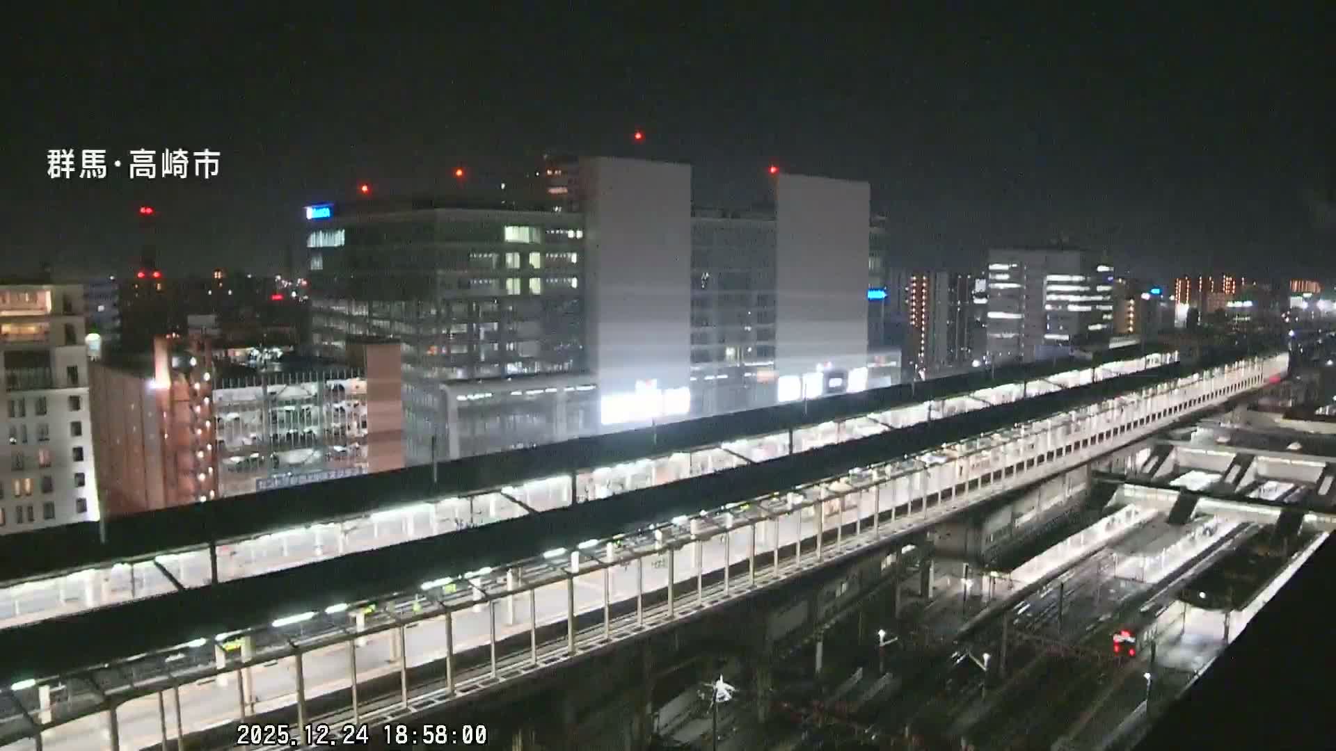 A nighttime aerial view reveals a sprawling city train station with elevated platforms and numerous tracks, flanked by illuminated buildings, all under a clear sky.