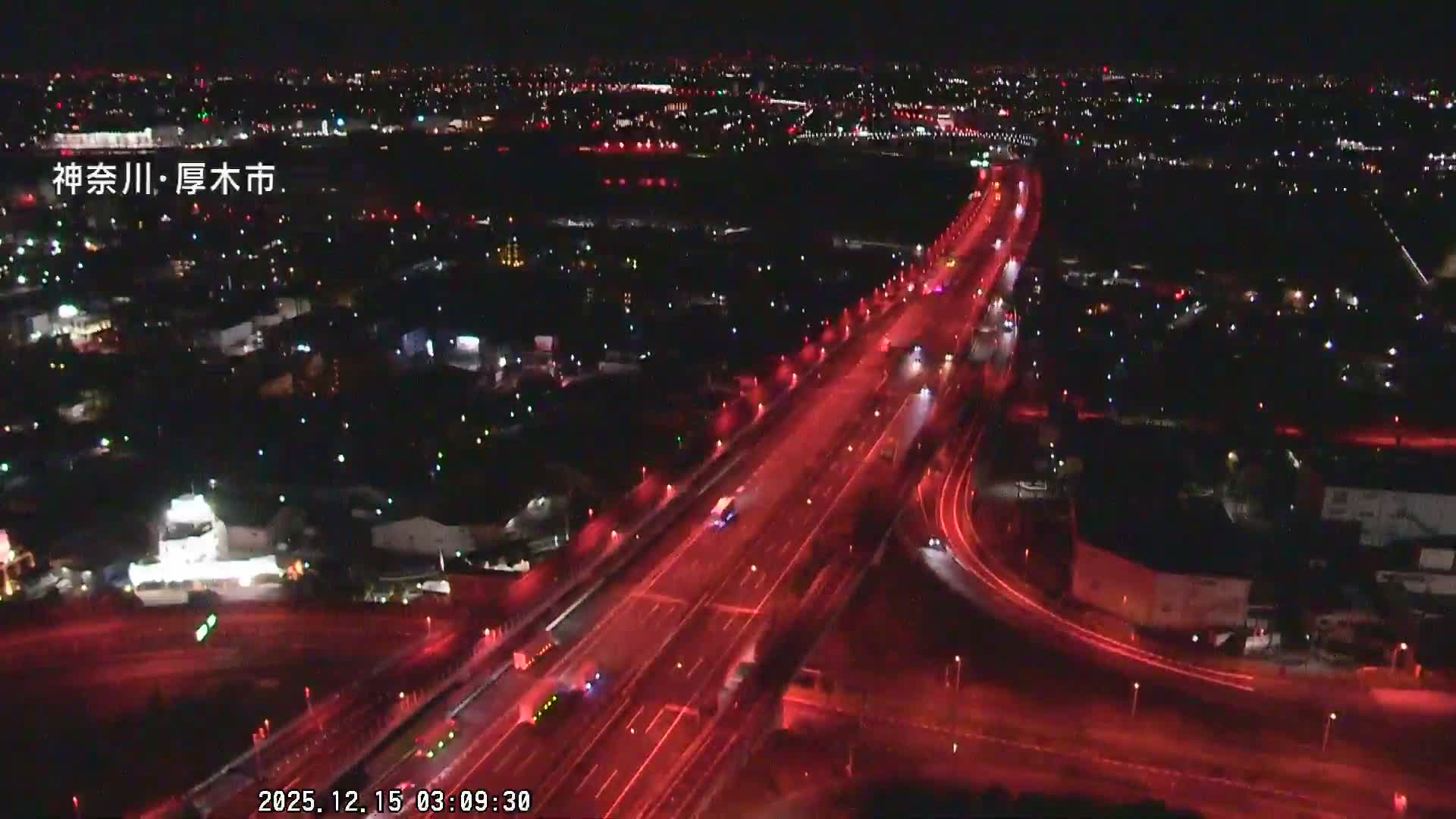 An aerial night view depicts a bustling city with prominent red-lit highways carrying vehicles, surrounded by numerous city lights fading into the distance under a clear night sky.