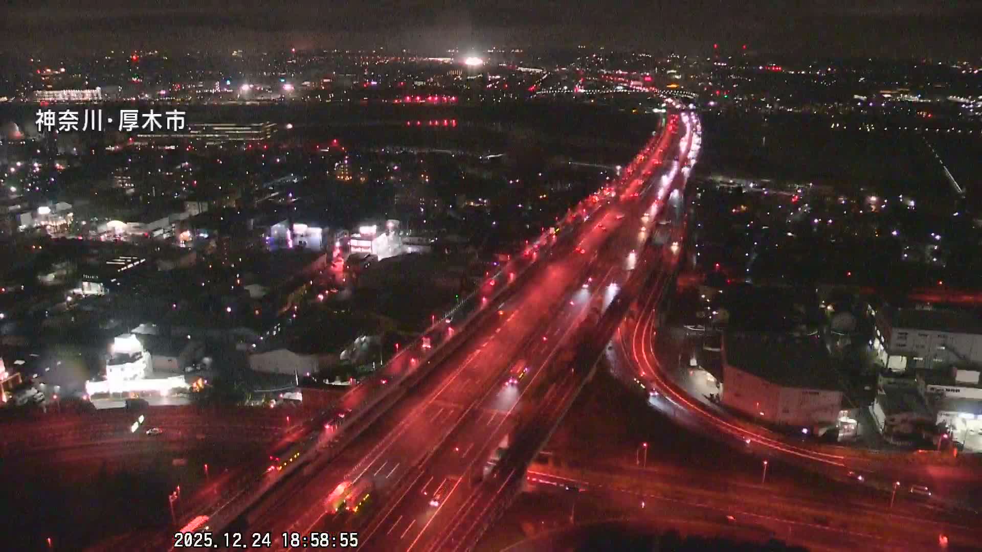 An aerial night view depicts a bustling city with prominent red-lit highways carrying vehicles, surrounded by numerous city lights fading into the distance under a clear night sky.