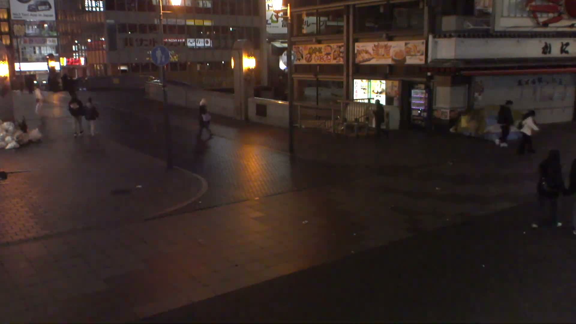 Pedestrians walk on wet city streets past brightly lit buildings and storefronts at night.