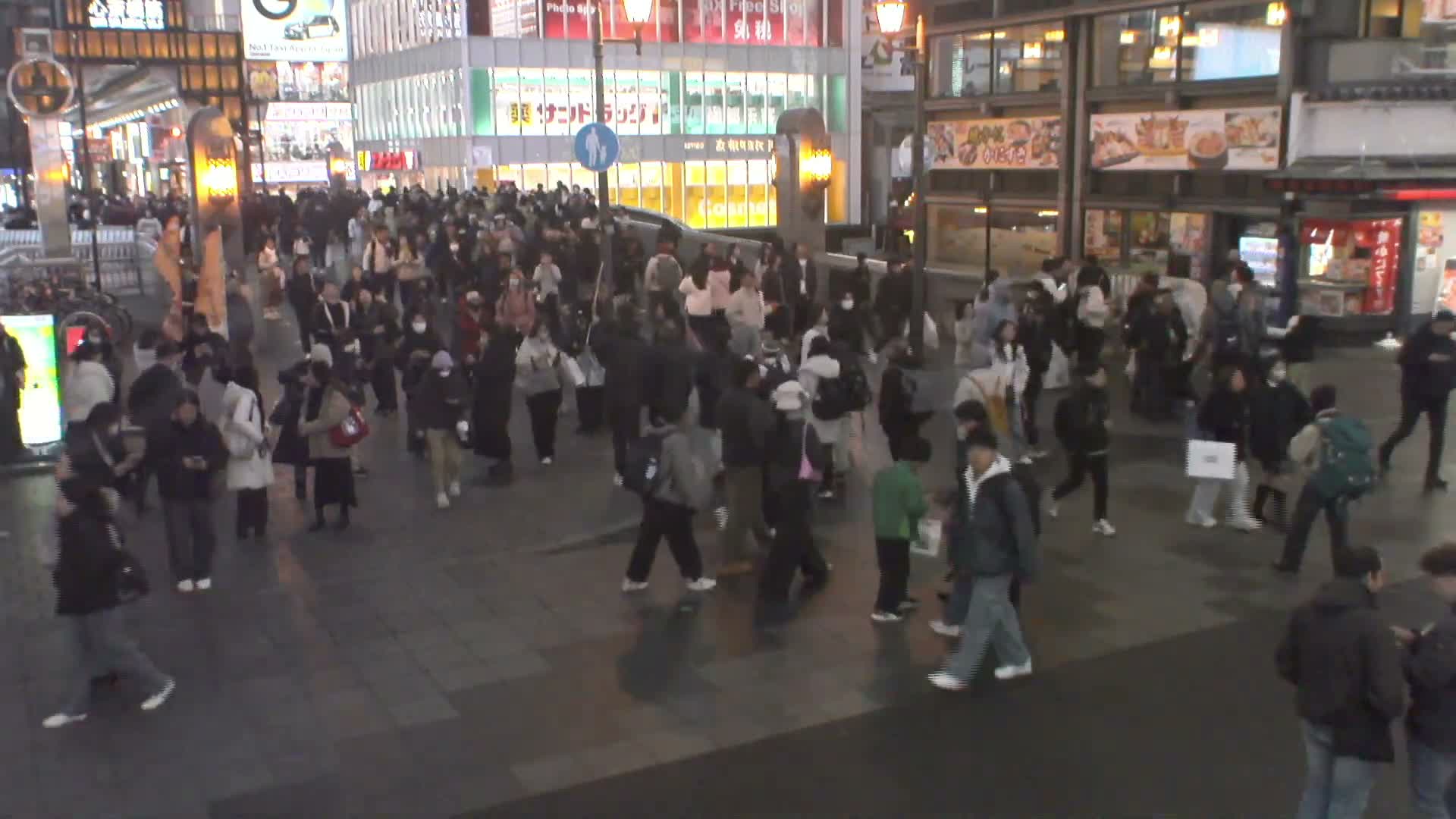A crowded city street is bustling with numerous pedestrians at night under dry, cool conditions, illuminated by vibrant storefront and street lights.