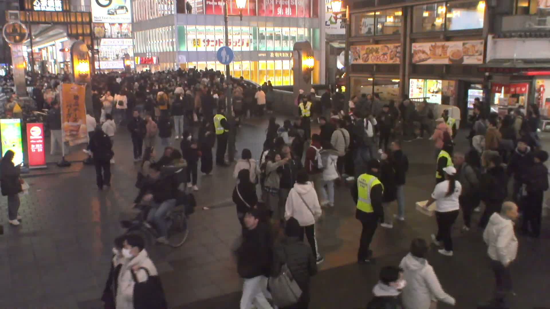 A bustling crowd of people mills about a brightly lit urban street at night, with illuminated buildings and a dark, reflective pavement under dry conditions.
