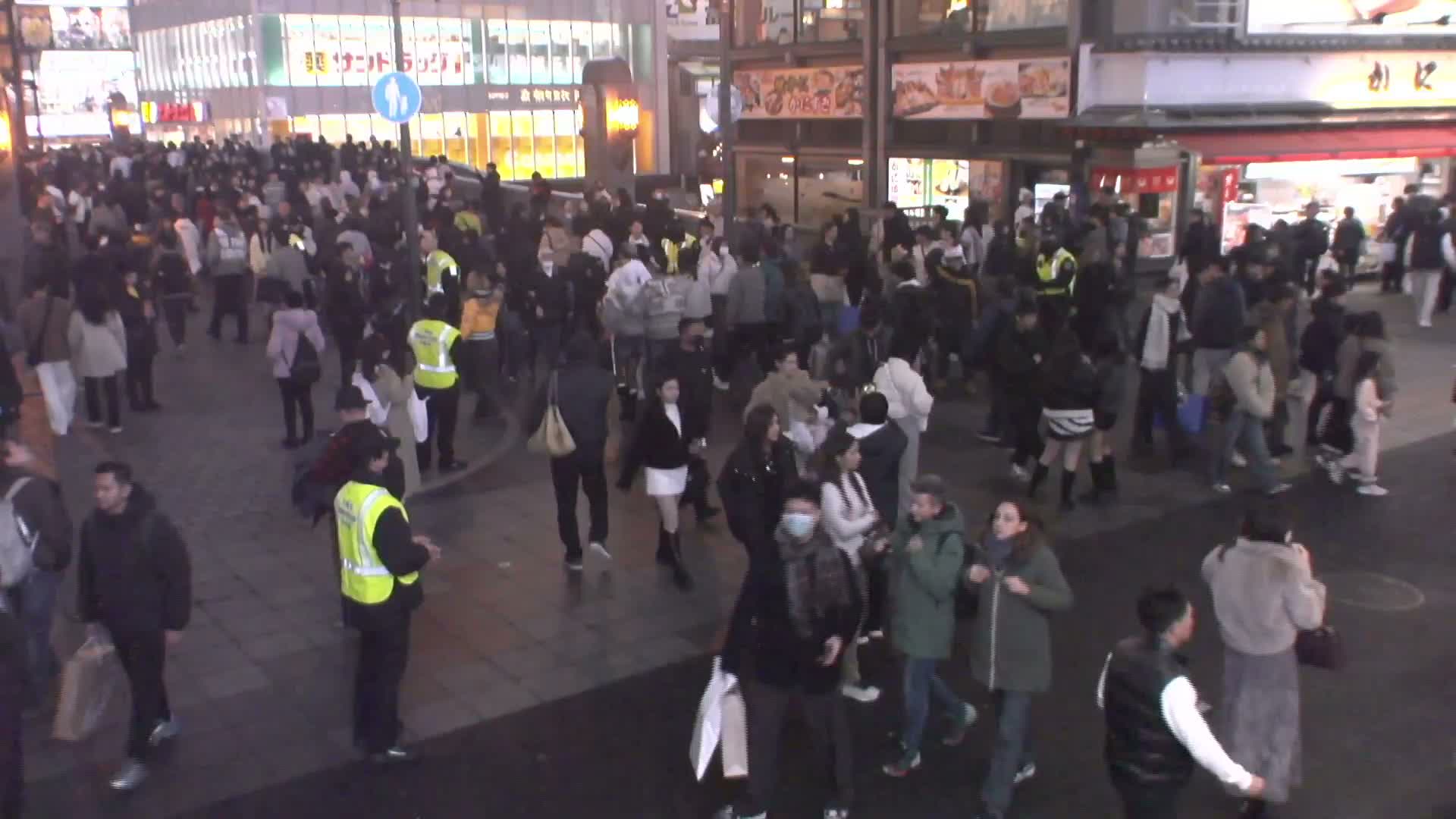 A bustling crowd of people mills about a brightly lit urban street at night, with illuminated buildings and a dark, reflective pavement under dry conditions.