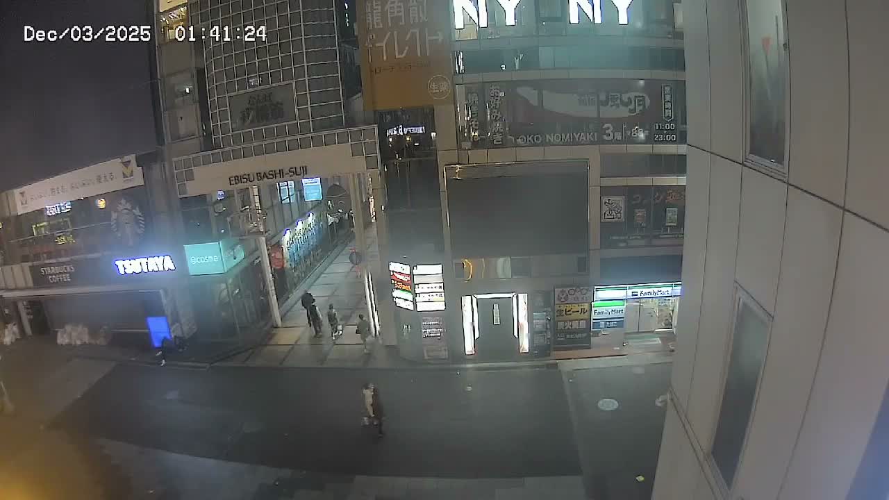 An elevated view captures a well-lit city street at night, with multiple buildings and storefronts illuminated by artificial lights as several pedestrians walk along the sidewalks under clear weather conditions.