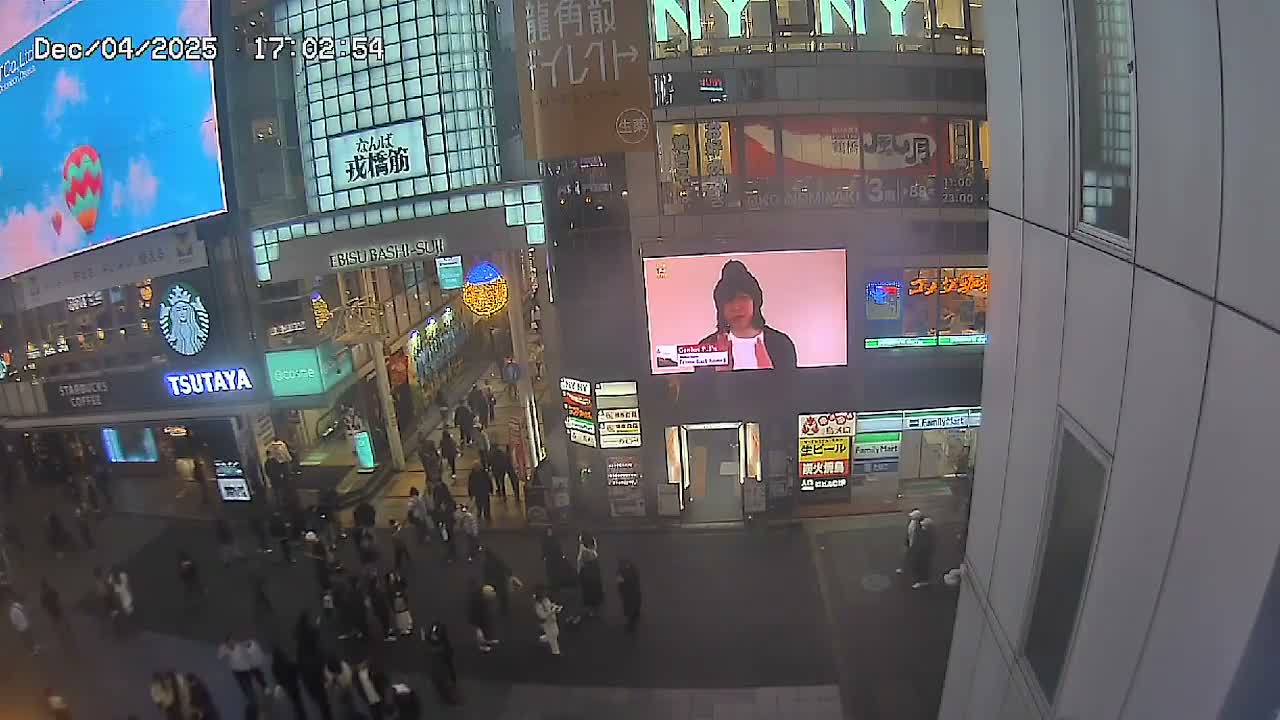 A crowded urban street scene at dusk is illuminated by the glowing screens of numerous large digital billboards and storefronts, with many pedestrians walking under clear, dry weather.