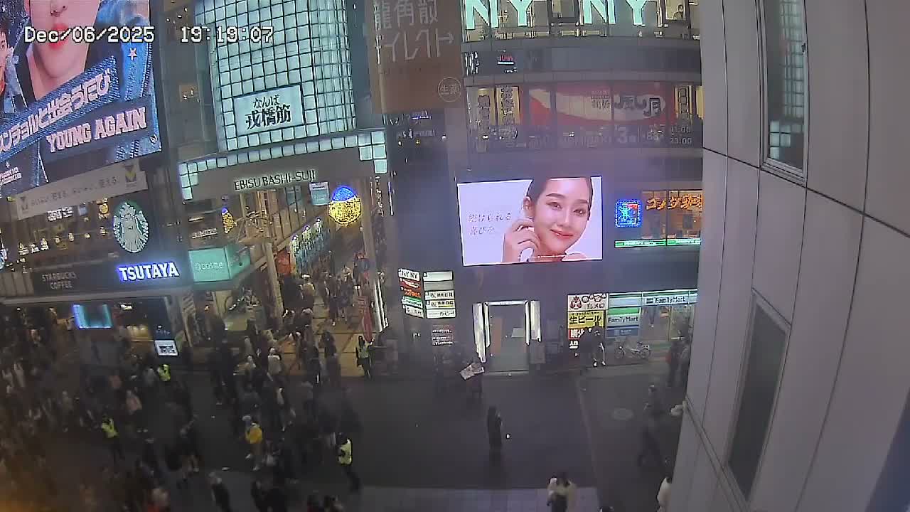 A bustling urban street is viewed from an elevated perspective at night, filled with numerous pedestrians beneath glowing digital billboards and illuminated storefronts.