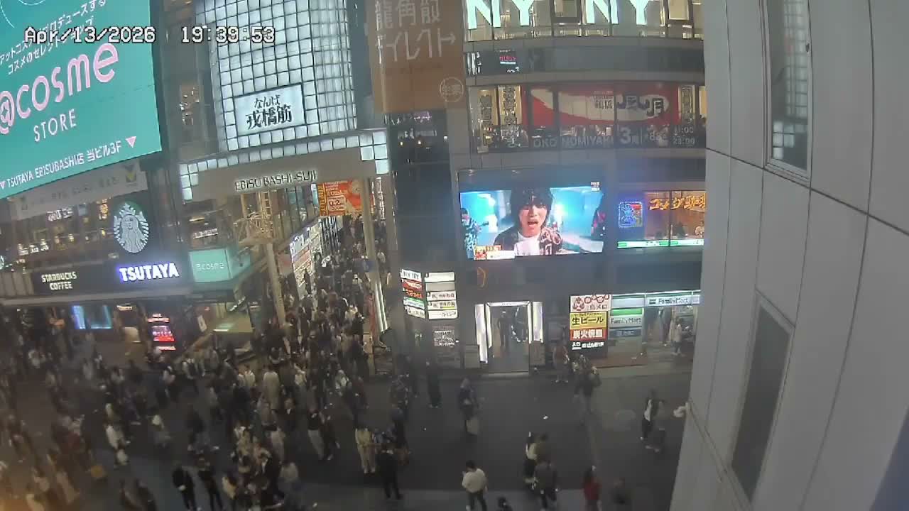 A bustling urban street is viewed from an elevated perspective at night, filled with numerous pedestrians beneath glowing digital billboards and illuminated storefronts.