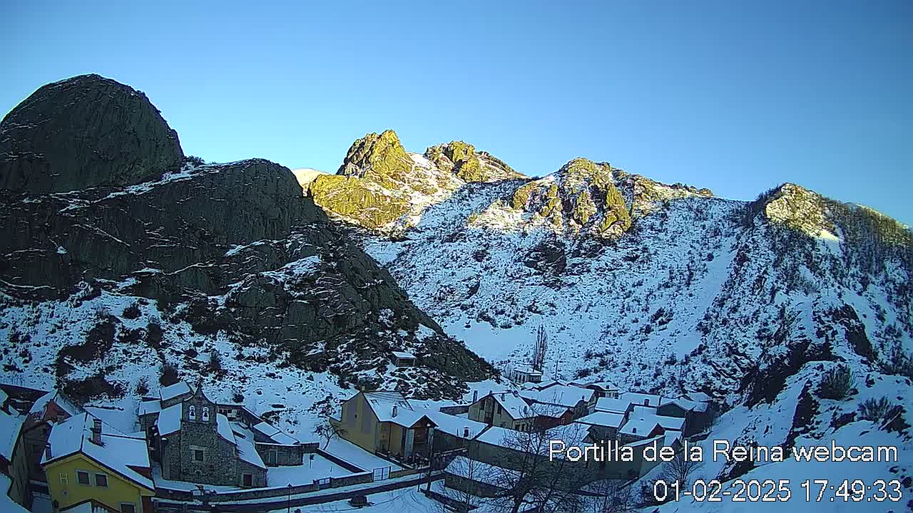 A snow-covered village nestled in a valley between steep, rocky mountains under a clear blue sky.