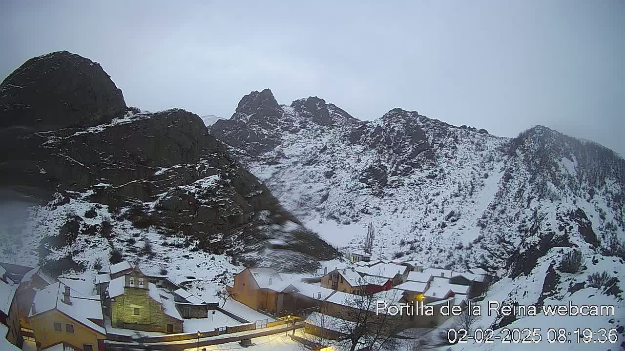 A snow-covered village nestled in a valley between dark, rocky mountains under an overcast sky.