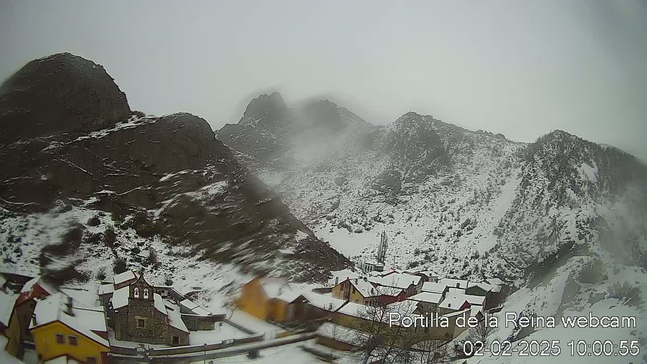 A snow-covered village nestled in a valley between snow-dusted, rocky mountains under an overcast sky.