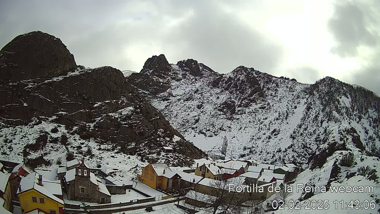 A snow-covered village nestled in a valley between dark, rocky mountains under a cloudy sky.