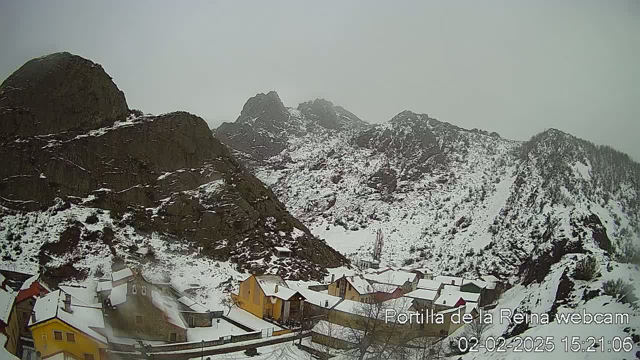 A snow-covered village nestled in a valley between rocky, snow-dusted mountains under an overcast sky.