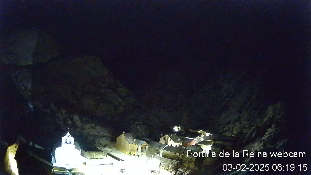 A small, snow-covered village is illuminated at night, nestled between dark, snow-dusted mountains under a clear, dark sky.