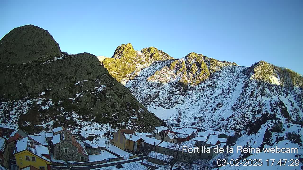 A snow-covered village nestled in a valley between steep, rocky mountains under a clear blue sky.