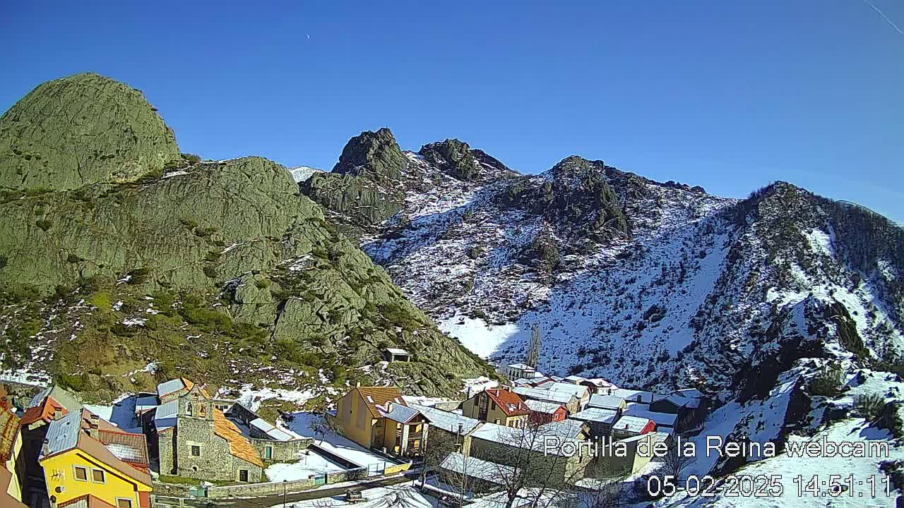 A small village nestled in a valley between snow-dusted, rocky mountains under a clear blue sky.