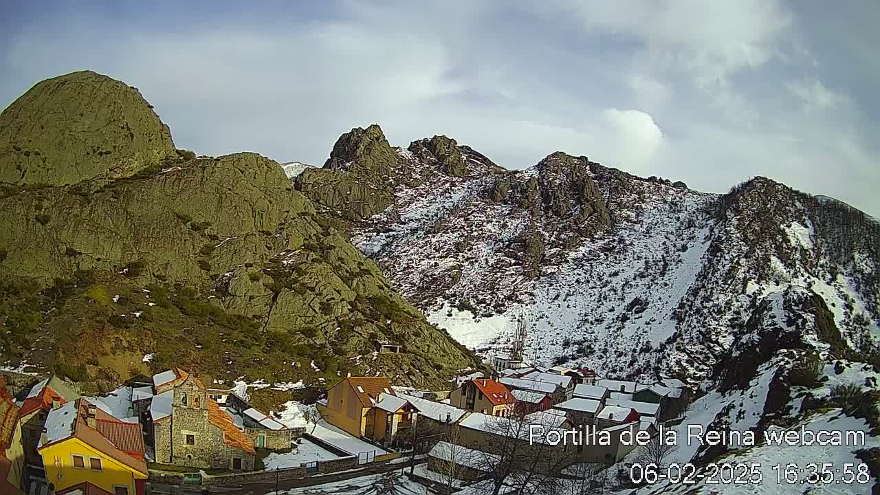 A small village nestled in a valley between snow-dusted, rocky mountains under a partly cloudy sky.