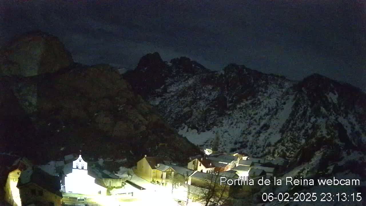 A small, snow-dusted village nestled in a dark, snow-capped mountain valley under a night sky.