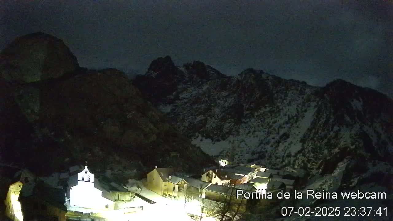 A nighttime view of a snow-dusted mountain range overlooking a small village illuminated with lights.