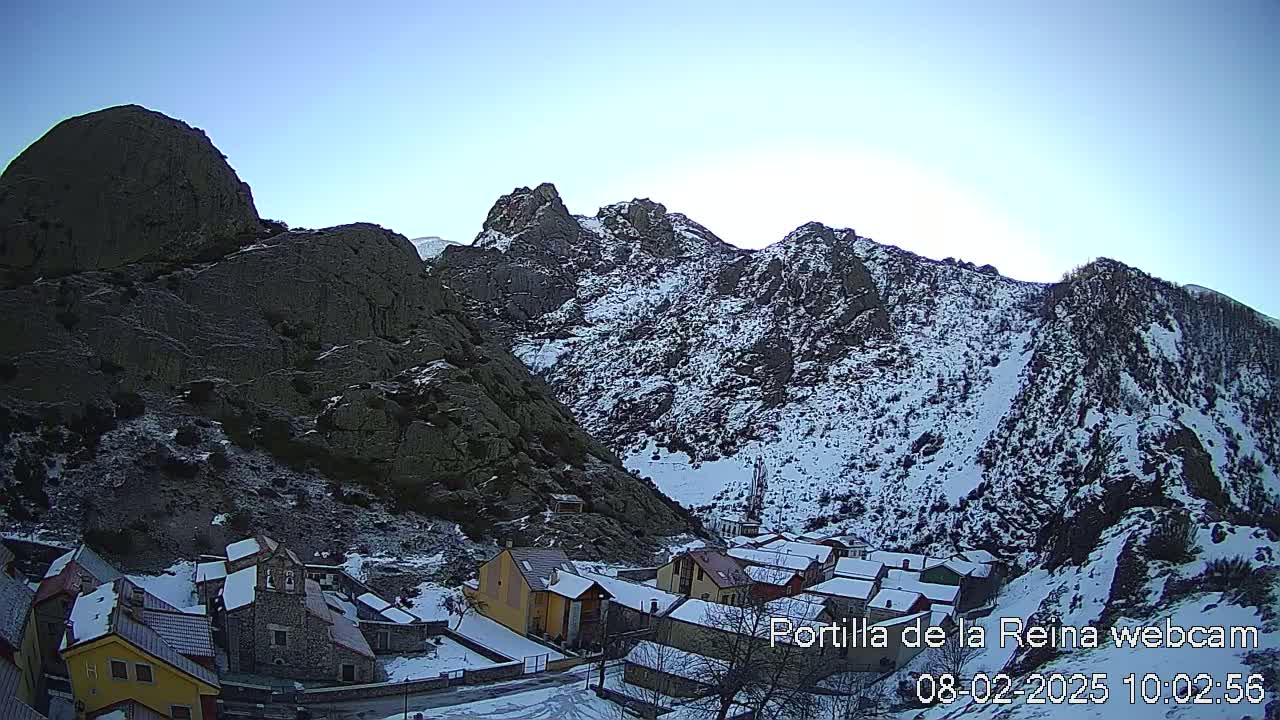 A snow-covered village nestled in a valley between steep, rocky mountains under a clear blue sky.
