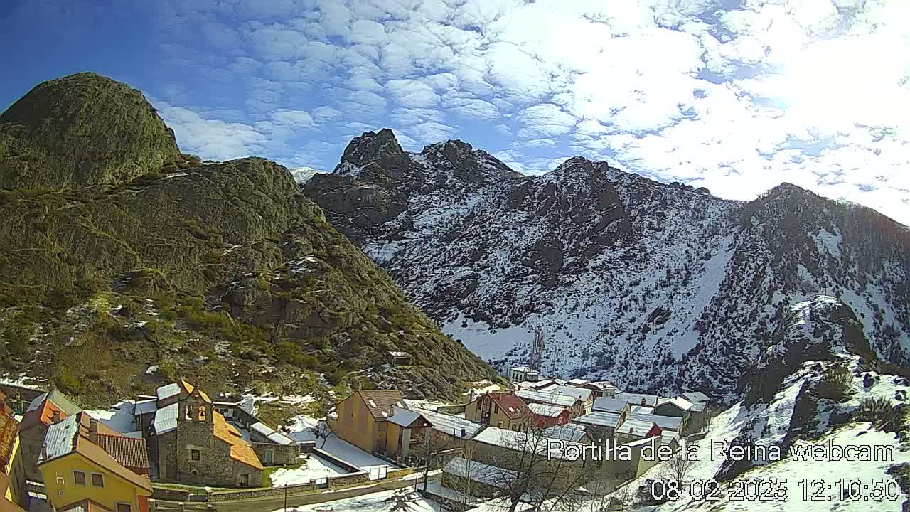 A small village nestled in a valley between snow-dusted mountains under a mostly sunny sky with scattered clouds.