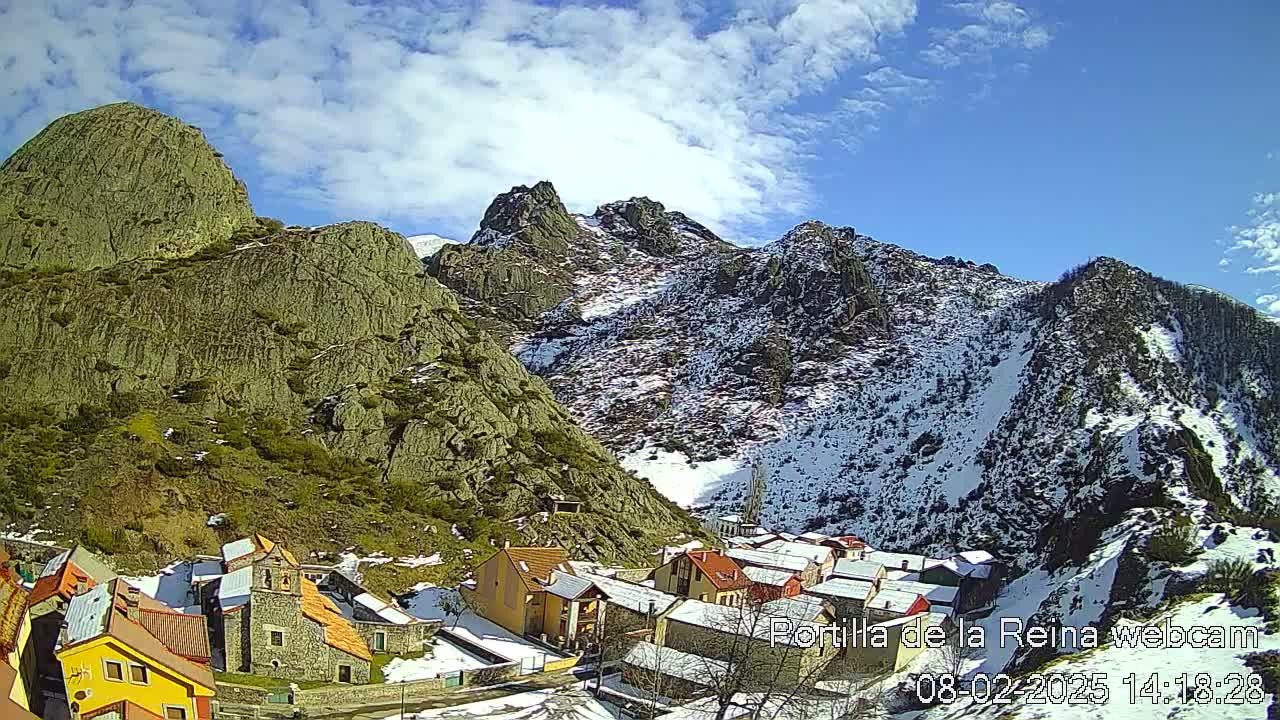 A small village nestled in a valley between snow-dusted mountains under a partly cloudy blue sky.