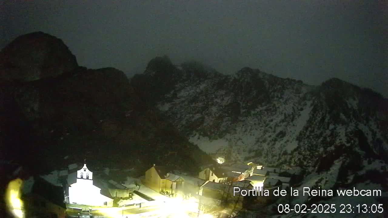 A nighttime view of a snow-dusted mountain village, illuminated by artificial lights, under a dark, overcast sky.