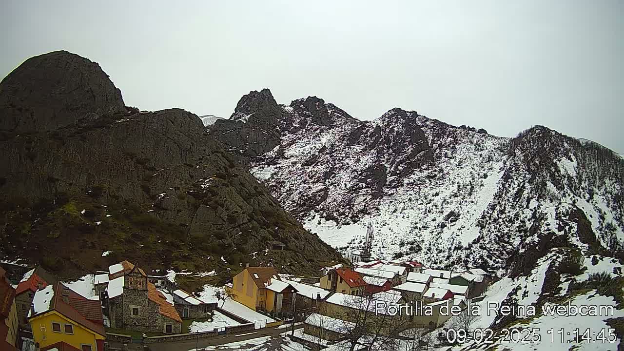 A small village nestled in a valley between snow-dusted, rocky mountains under an overcast sky.