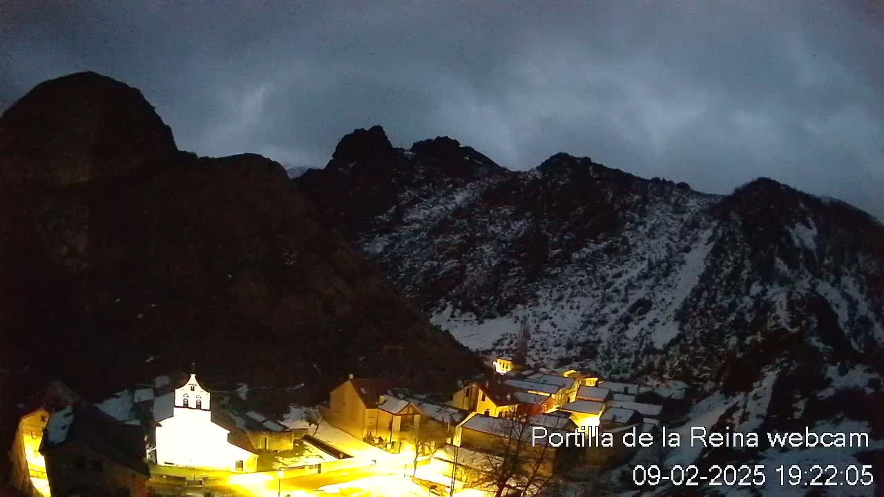 A small, illuminated village nestled in a snow-dusted mountain valley under a dark, cloudy sky.
