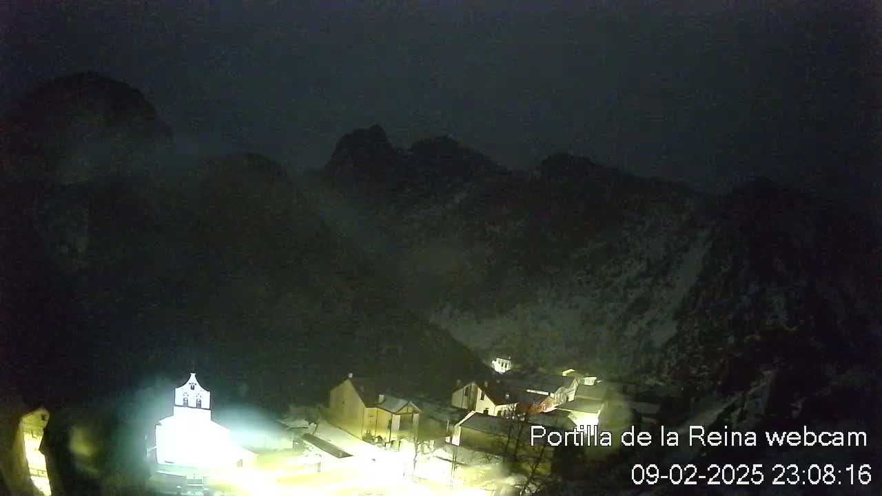 A nighttime view of a small village nestled in a valley surrounded by dark mountains under a cloudy sky.