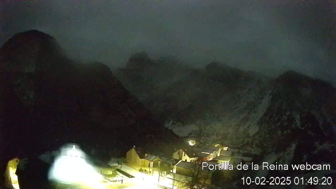 A small, snow-covered village nestled in a valley between dark, foggy mountains at night.