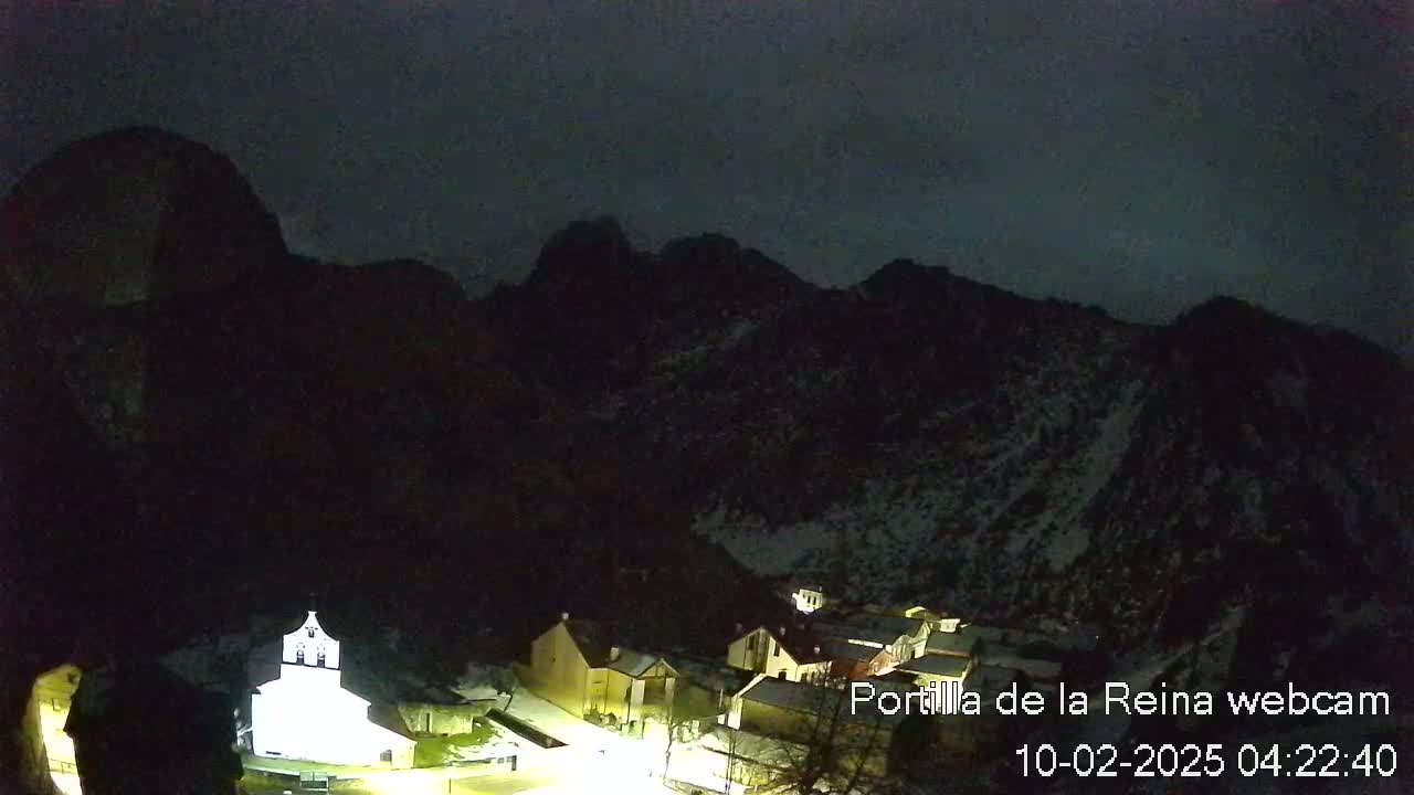 A nighttime view of a small village nestled in a snow-dusted mountain valley under a dark, cloudy sky.