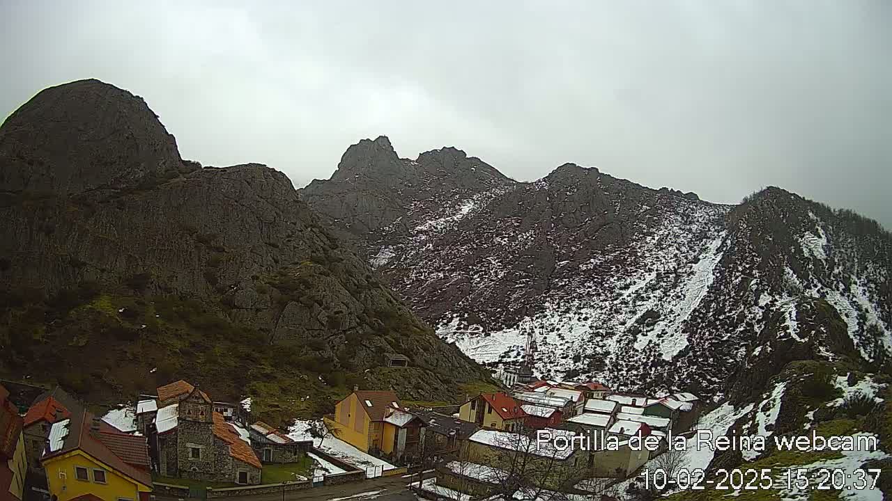 A small village nestled in a valley between snow-dusted, rocky mountains under an overcast sky.