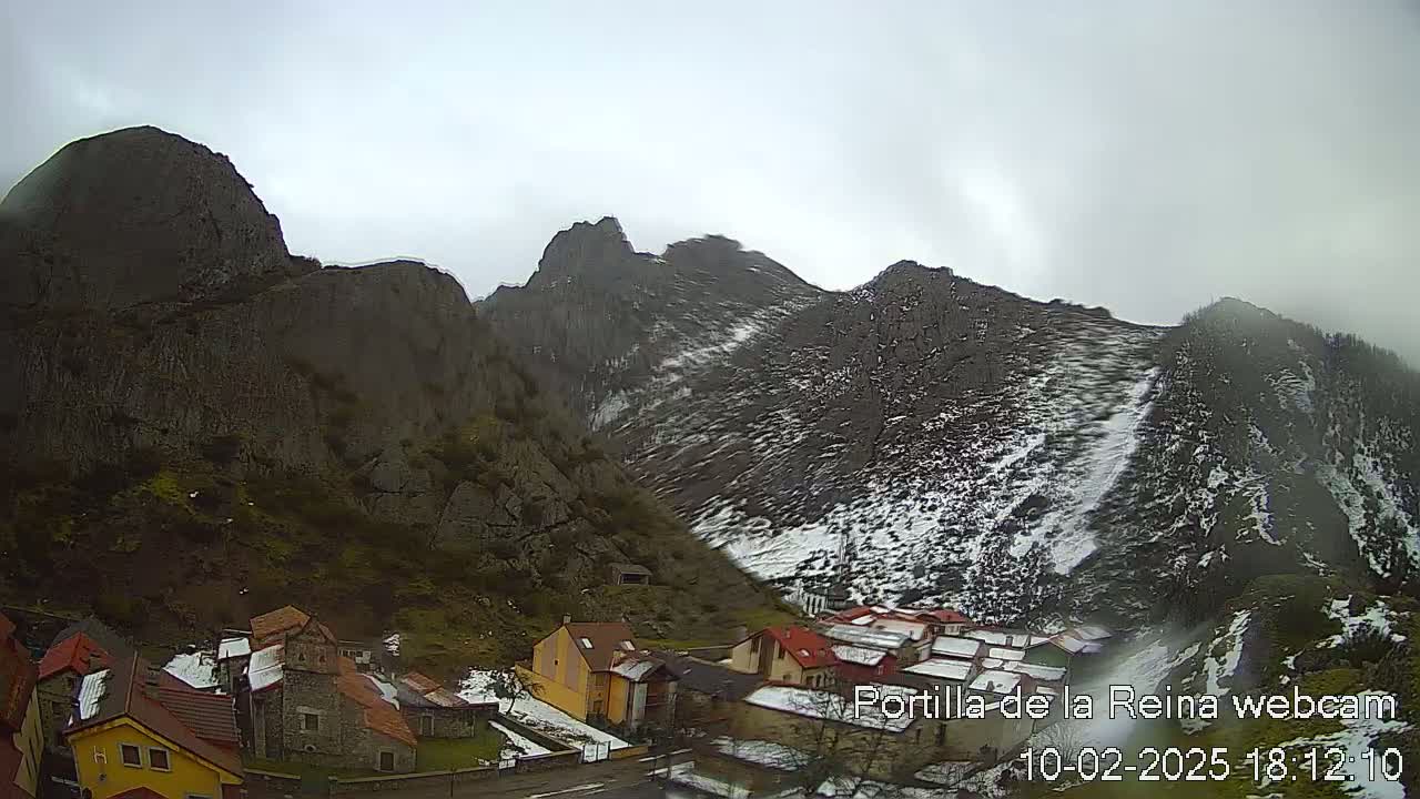 A small village nestled in a valley between snow-dusted, rocky mountains under an overcast sky.
