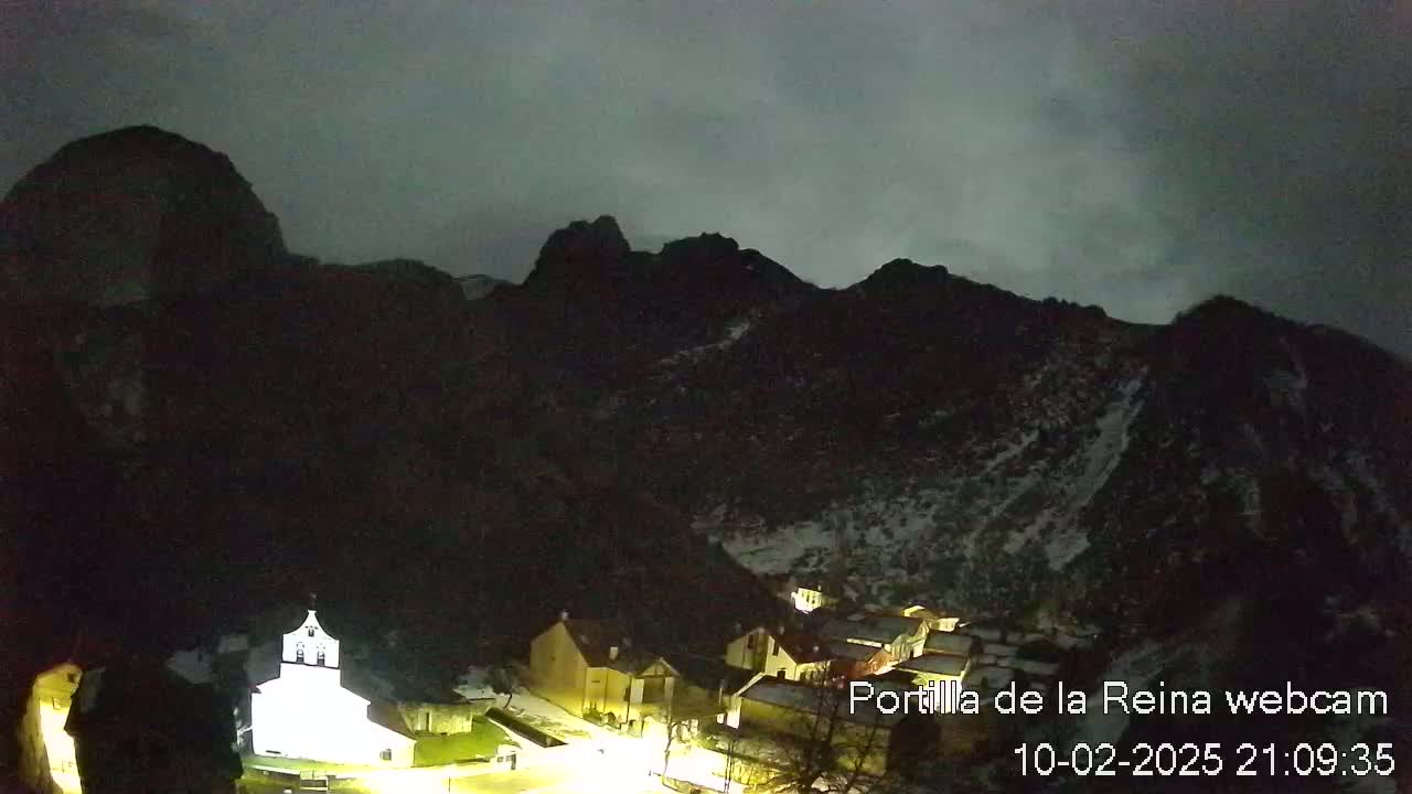 A small village nestled in a valley below dark, snow-dusted mountains under a cloudy night sky.