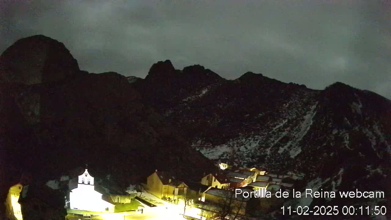 A small, illuminated village nestled in a dark, snow-dusted mountain valley under a cloudy night sky.