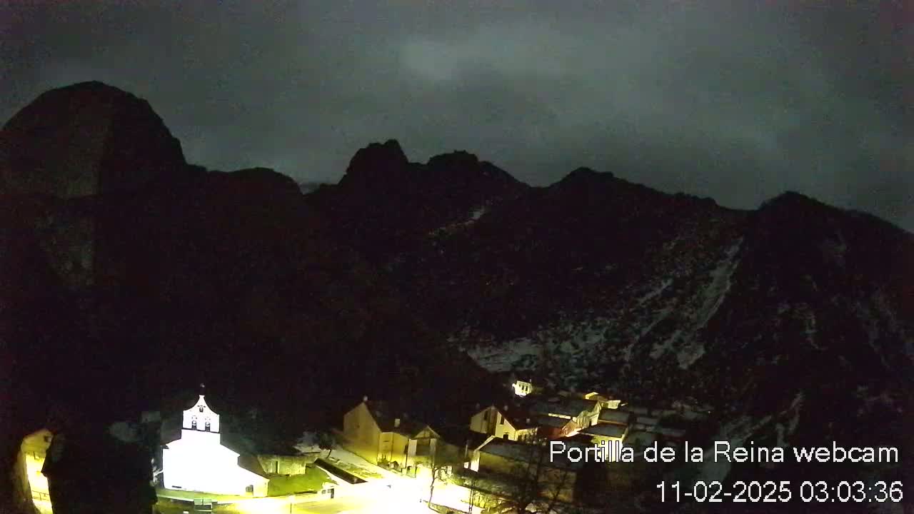 A small village nestled in a valley between dark, snow-dusted mountains under a cloudy night sky.