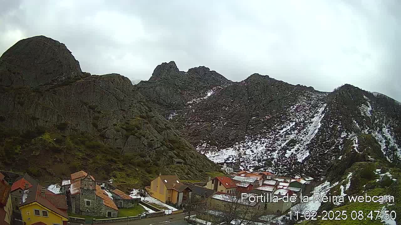 A small village nestled at the base of snow-dusted, rocky mountains under a cloudy sky.