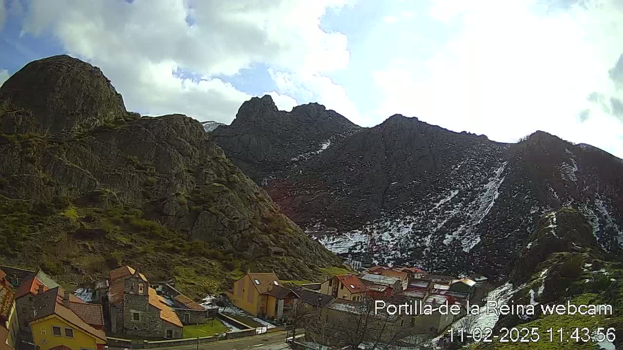 A small village nestled at the base of dark, rocky mountains partially covered in snow under a partly cloudy sky.
